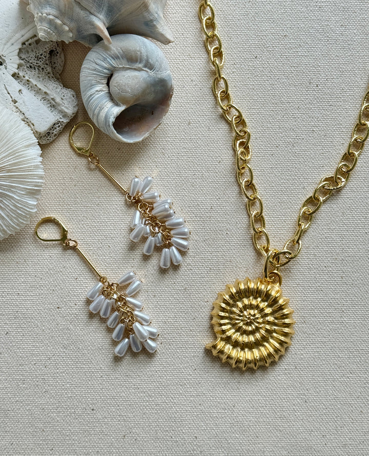 A gold-colored necklace with a conch shell pendant, accompanied by matching earrings, displayed on a beige background with shells and a chain.