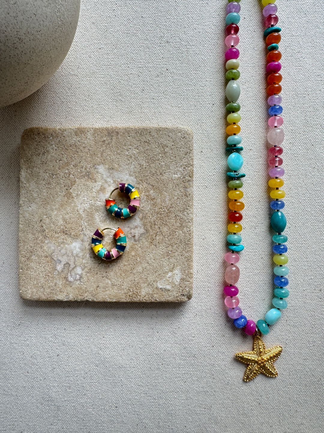 A multicolored beaded necklace with a gold starfish pendant, accompanied by a matching pair of earrings, displayed on a stone surface.