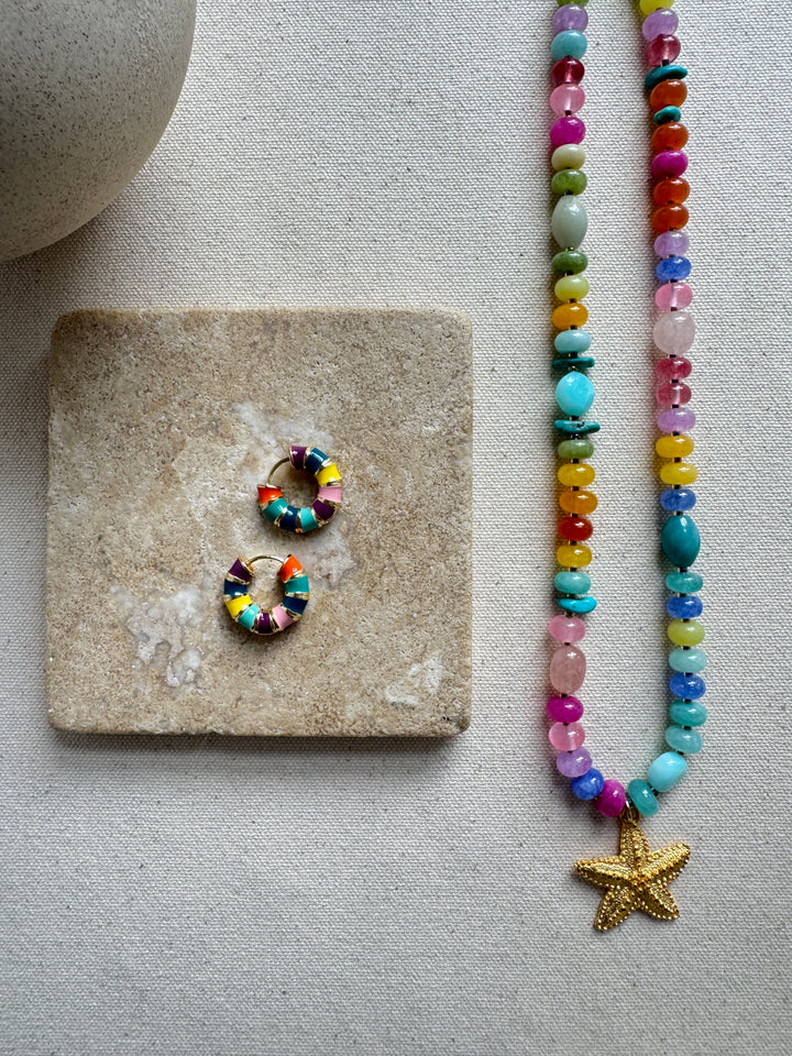 A multicolored beaded necklace with a gold starfish pendant, accompanied by a matching pair of earrings, displayed on a stone surface.