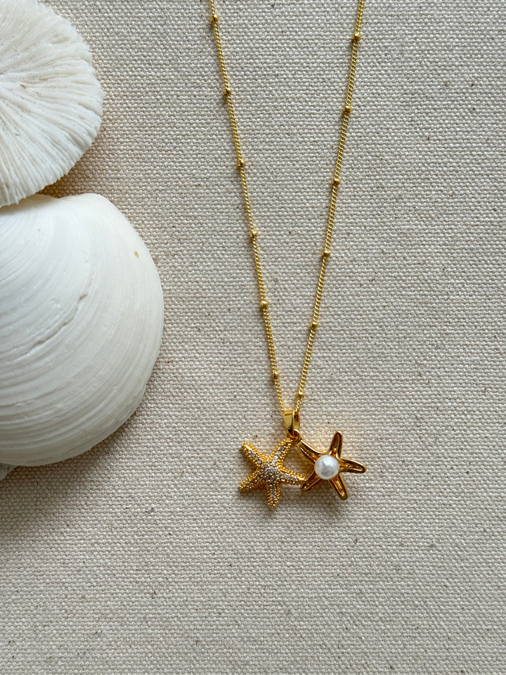 A gold-filled satellite chain necklace with two textured starfish charms and a pearl, displayed on a beige fabric surface alongside a conch shell.
