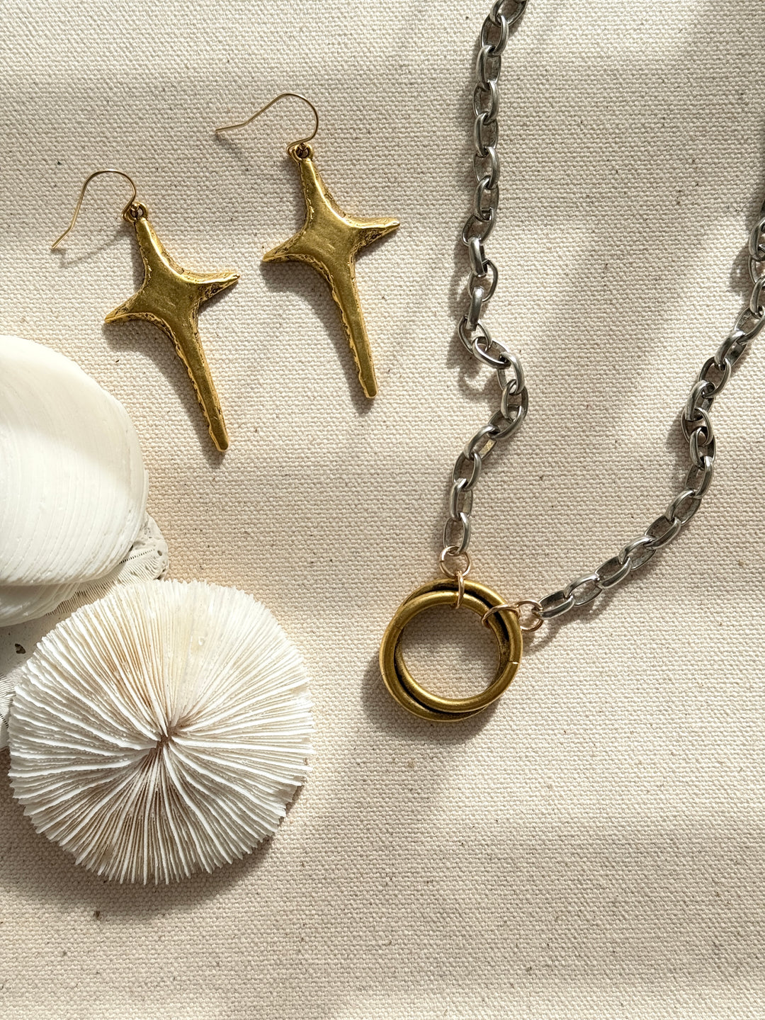 A gold  and silver necklace with a circular pendant, along with a pair of gold-colored star earrings , displayed on a textured background with a white shell and a ring.