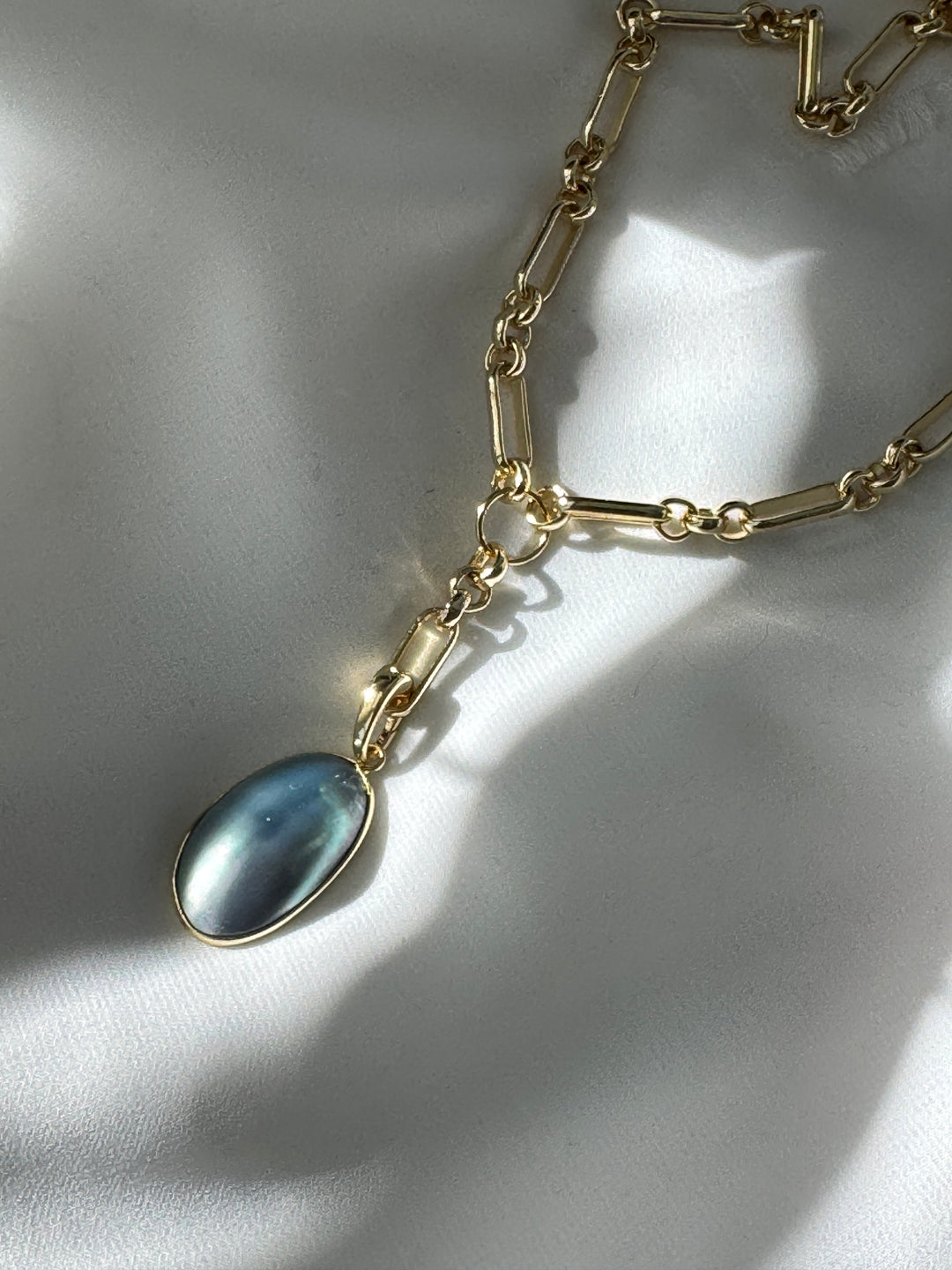 A gold-filled necklace with a long and short chain featuring a freeform blue pearl removable pendant with a spring clasp, displayed on a white fabric surface.