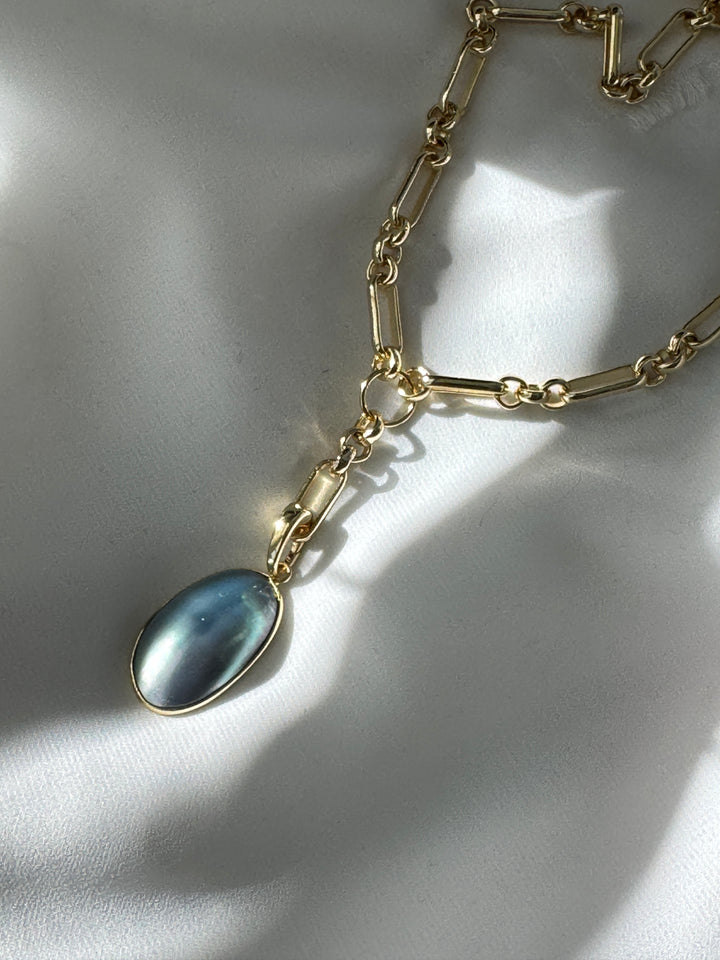 A gold-filled necklace with a long and short chain featuring a freeform blue pearl removable pendant with a spring clasp, displayed on a white fabric surface.