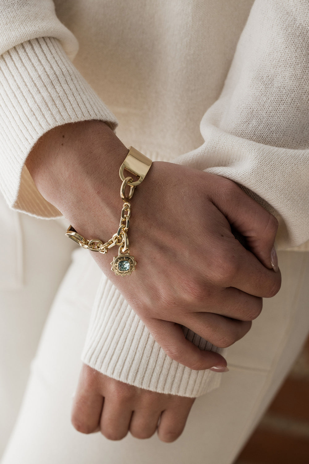 A person wearing a white sweater showcasing a gold-colored bangle bracelet with a sunburst charm and an aqua green crystal on the wrist.