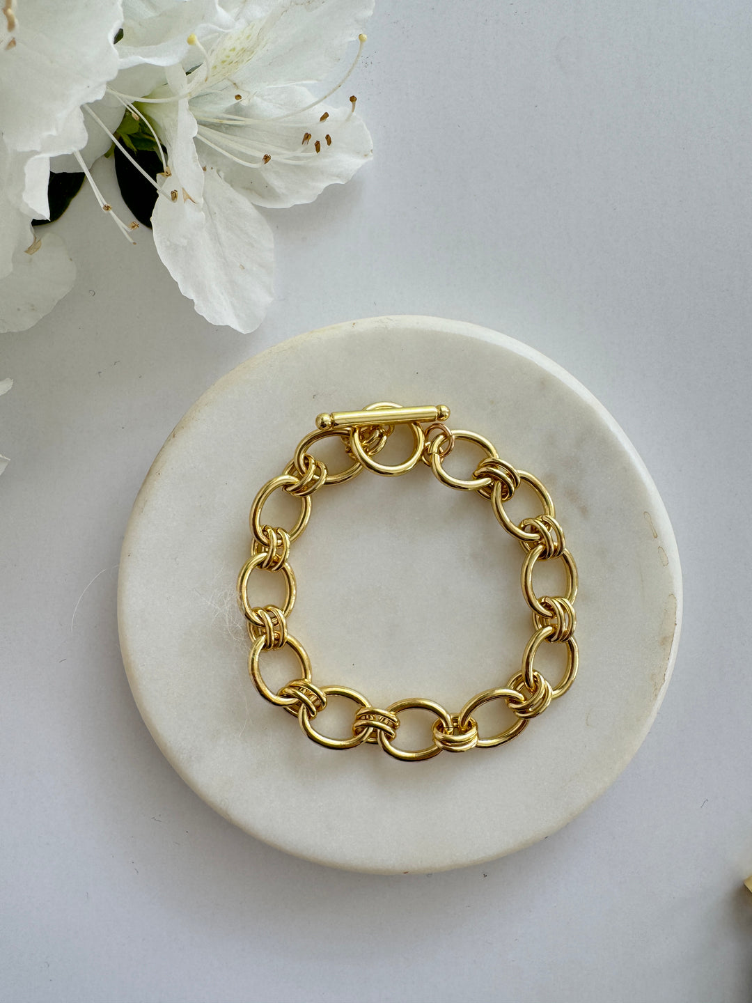 A gold-filled bracelet with double links and a toggle clasp, displayed on a white marble plate next to white flowers.